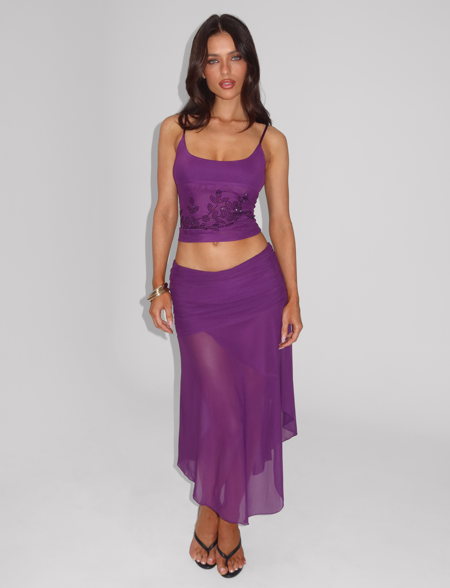 A woman with long brown hair wears a purple sleeveless crop top and the TIGERMIST MARIANA SKIRT - PURPLE featuring an asymmetric wrap design, standing against a plain white background.