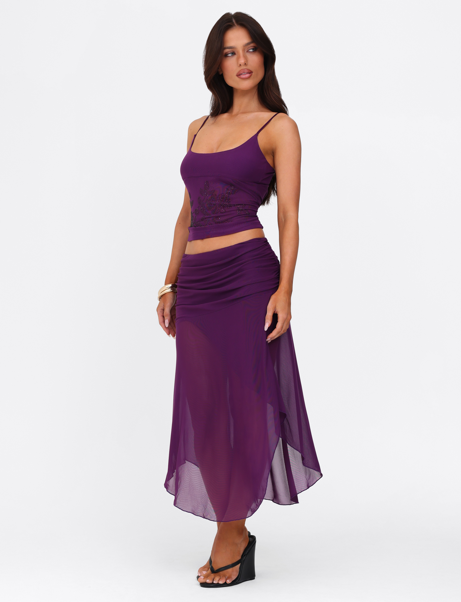 A woman models the TIGERMIST MARIANA SKIRT - PURPLE with a sleeveless top and heels, posing against a white backdrop. She stands with one hand on her hip, highlighting the skirt’s elegant purple shade.