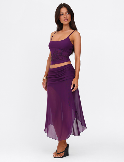 A woman models the TIGERMIST MARIANA SKIRT - PURPLE with a sleeveless top and heels, posing against a white backdrop. She stands with one hand on her hip, highlighting the skirt’s elegant purple shade.