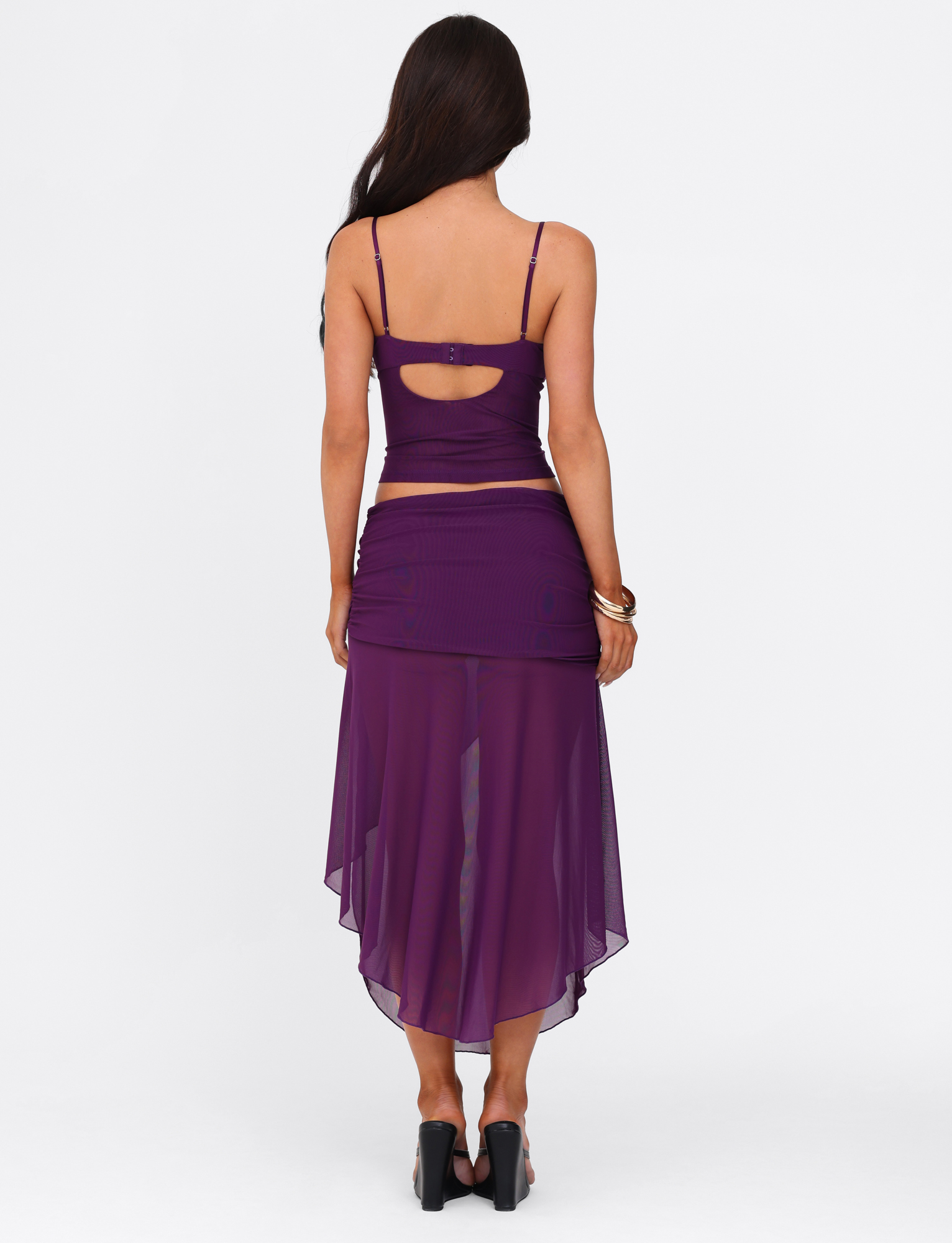 A woman with long brown hair wears a sleeveless purple crop top and the TIGERMIST MARIANA SKIRT - PURPLE, featuring a sheer asymmetric wrap design, paired with black heels, standing back to the camera against a white background.