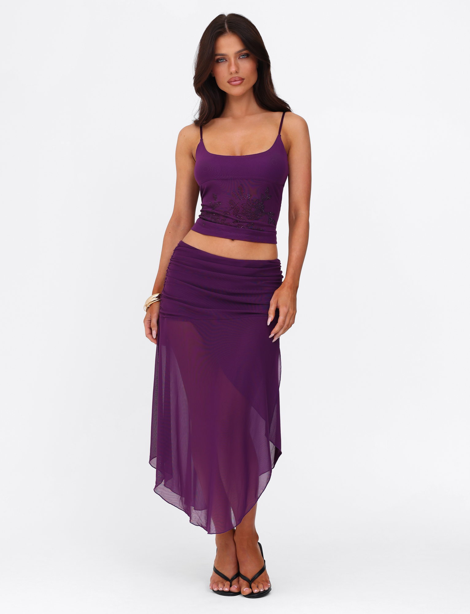 A woman with long dark hair wears a sleeveless purple crop top and the TIGERMIST MARIANA SKIRT - PURPLE in an asymmetric wrap style, paired with black strappy heels, against a plain white background.