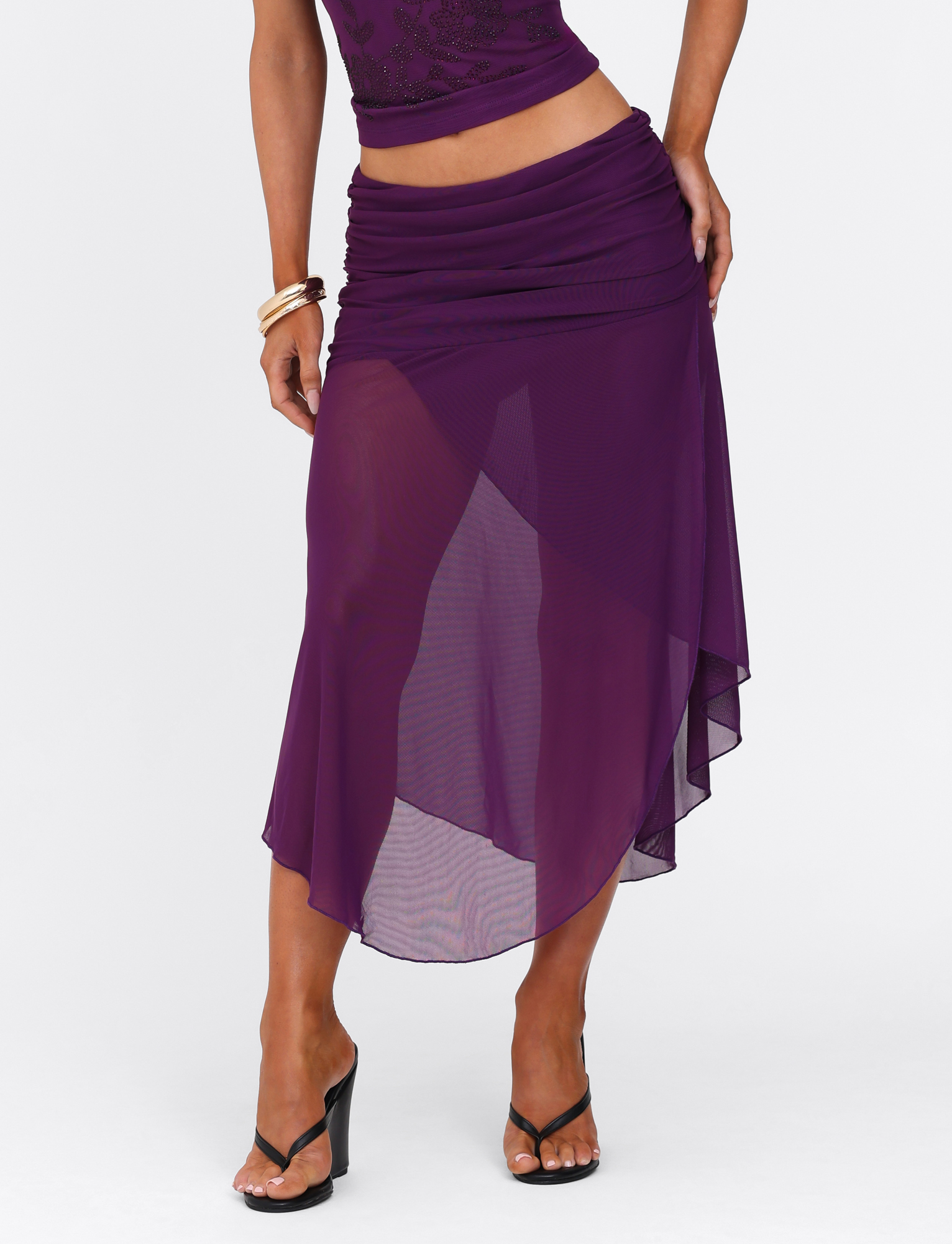 A person stands against a plain white background wearing the TIGERMIST MARIANA SKIRT - PURPLE, styled with its matching top, high-heeled black sandals, and gold bangles.