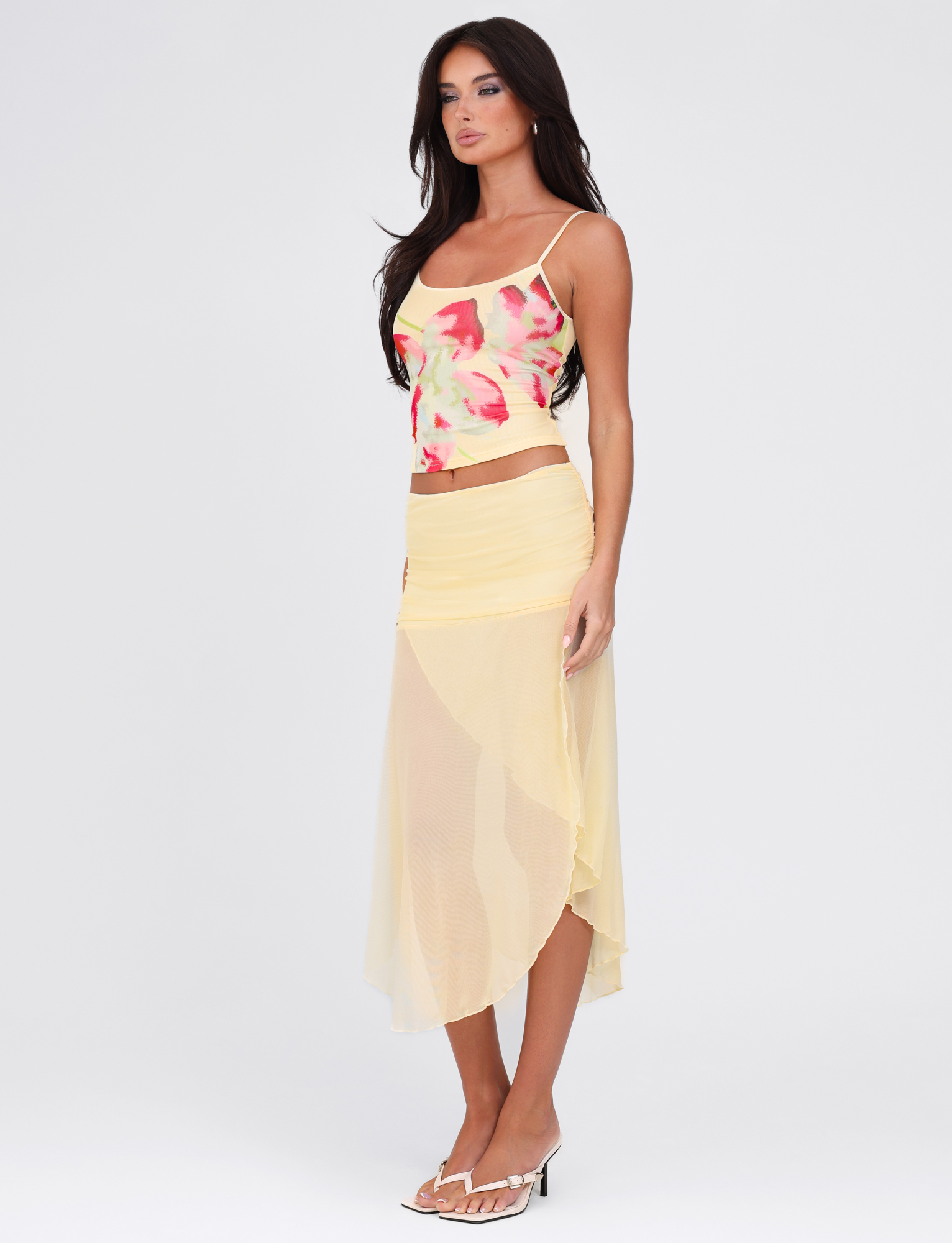 A woman with long dark hair wears a colorful tank, the TIGERMIST MARIANA SKIRT in pale yellow mesh, and white heeled sandals while posing against a plain white background.