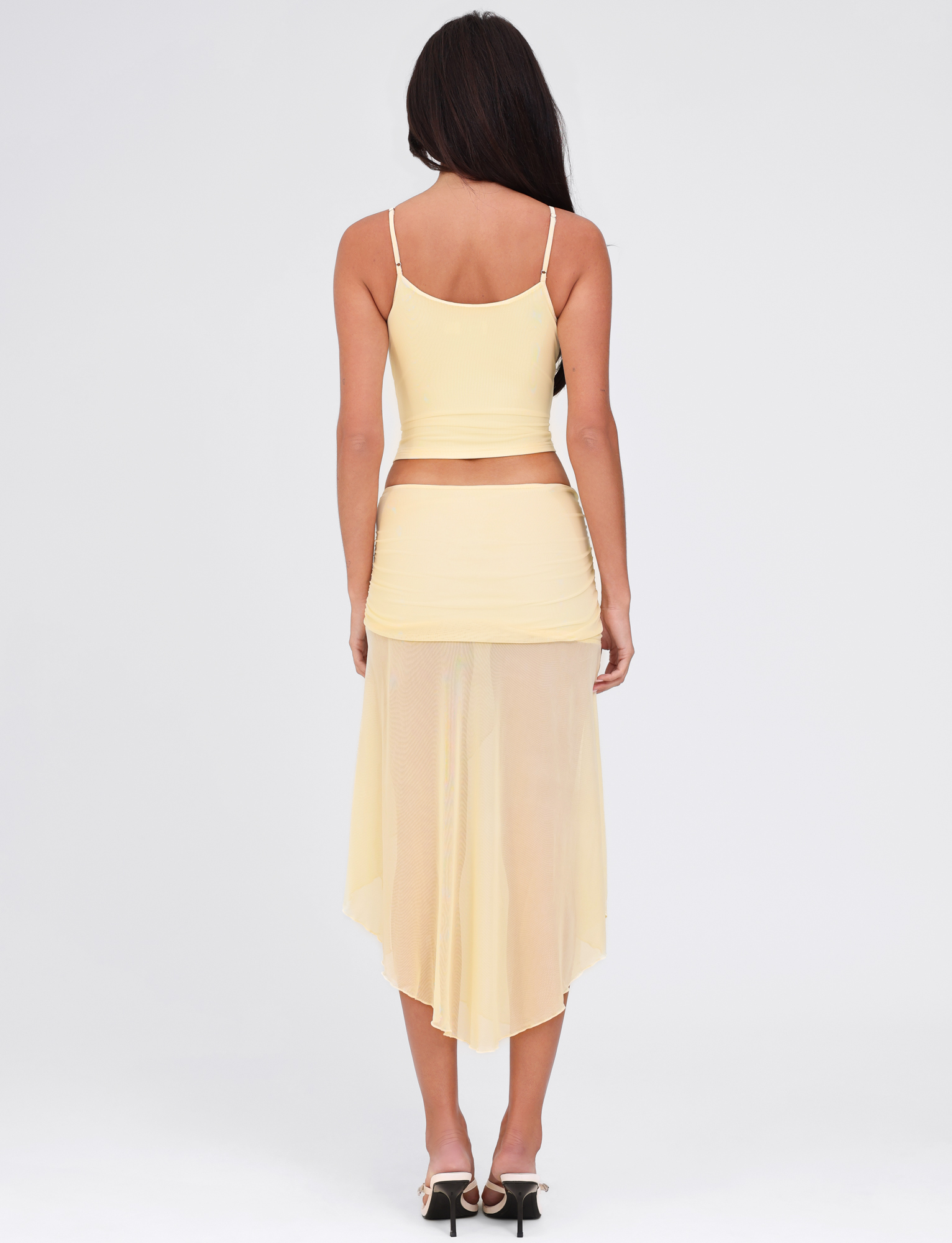 A woman with long dark hair, seen from behind, wears the TIGERMIST MARIANA SKIRT in pale yellow with a matching sleeveless crop top and high-heeled sandals, standing against a plain white background.