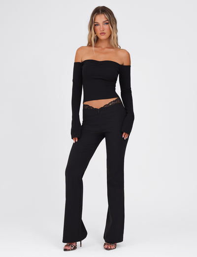 A woman dressed in the NYOMI PANT in black by TIGERMIST stands against a plain white background, wearing a fitted black off-shoulder top with flared leg pants adorned with black lace detail and open-toe heels.