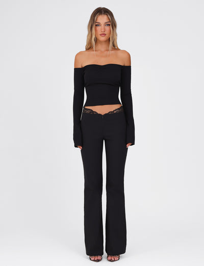 A woman stands against a plain white background, wearing a black, off-the-shoulder top with long sleeves paired with the NYOMI PANT - BLACK from TIGERMIST, featuring high stretch fabric and lace details. Her long, wavy hair cascades down as she completes the look with stylish sandals.