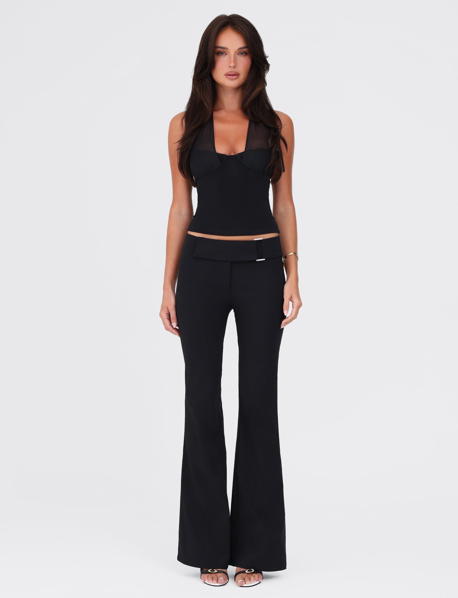 A woman with long, dark hair stands against a plain white background. She is wearing a black sleeveless top and the VESPER PANT - BLACK by TIGERMIST, featuring a fitted silhouette that accentuates her low-rise waist. With a neutral expression, her arms rest relaxed by her sides. She completes the outfit with black heeled sandals.