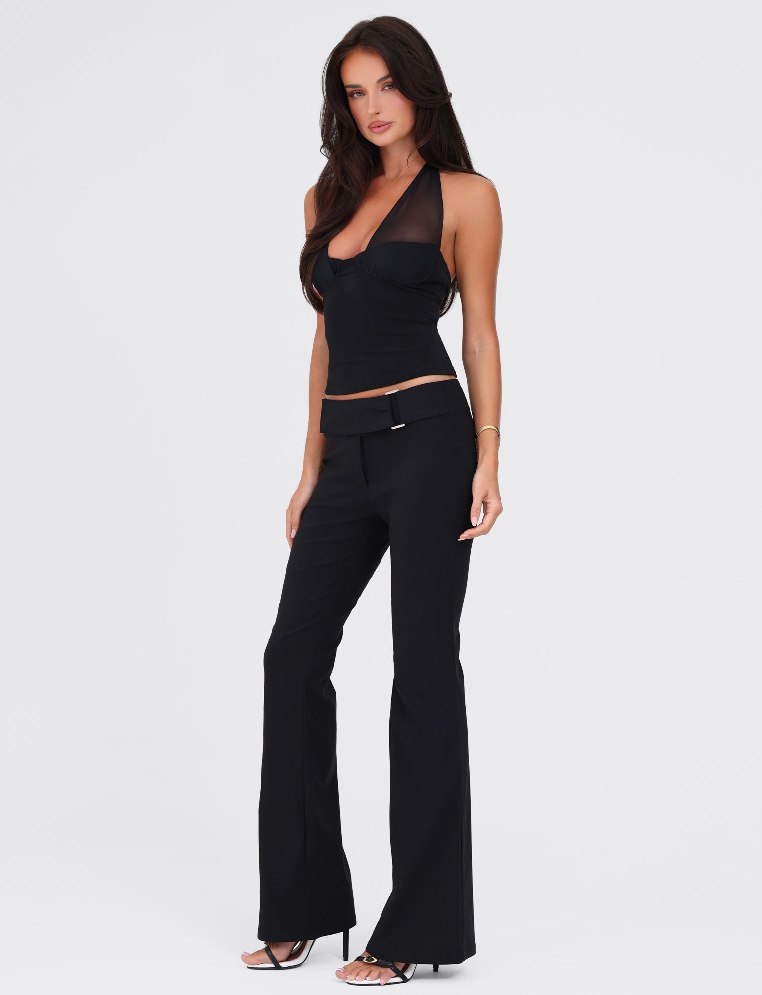 A woman with long dark hair stands in a confident pose, wearing a fitted black sleeveless halter top and the TIGERMIST VESPER PANT - BLACK, which has a low-rise waist and flared legs. She accessorizes with a single bracelet on her left wrist and black open-toe heels. She stands against a plain white background.
