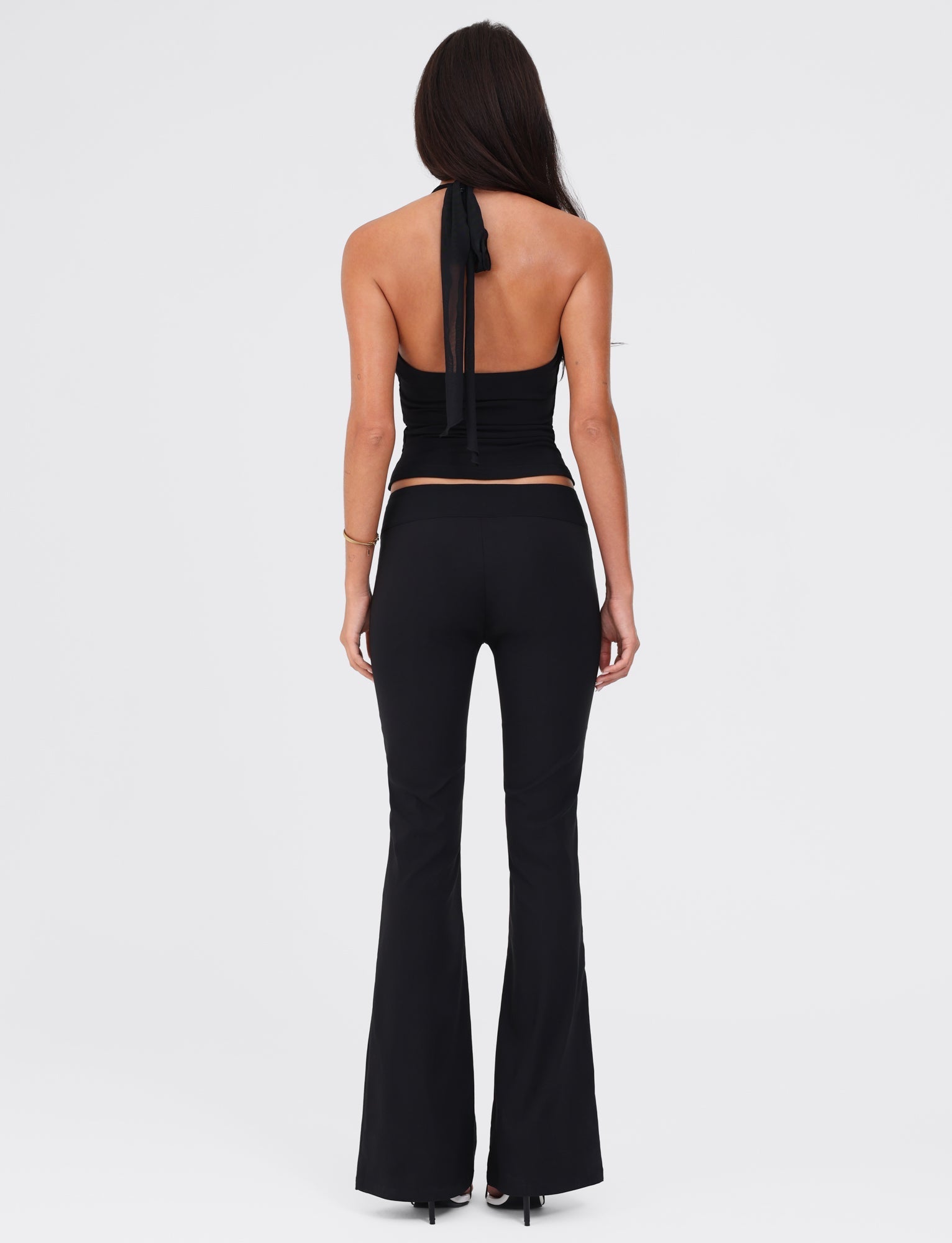 A person with long brown hair stands with their back to the camera, dressed in a black strapless top that ties at the back and paired with TIGERMIST's VESPER PANT - BLACK, which exhibits a fitted silhouette and low-rise waist. The plain white background highlights their black heeled shoes.