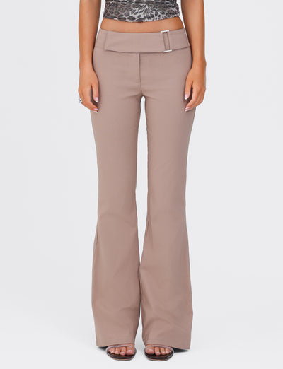 A person wearing the VESPER PANT in Brown Tan by TIGERMIST, featuring flared legs, a high waist, and a belt buckle detail. Paired with an animal print top, the individual stands with their hands relaxed at their sides, exuding a polished yet casual style.