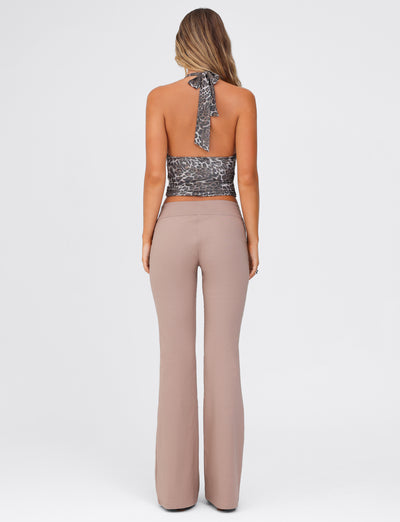 A woman is seen from the back, with long wavy hair. She is wearing a leopard print halter top that ties at the neck and TIGERMIST's VESPER PANT in brown and tan, which are high-waisted and flared. The background is plain and light grey.