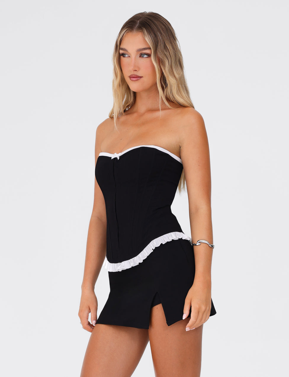 A woman with long, wavy blonde hair is wearing the TIGERMIST MILLIE CORSET 2.0 - BLACK, featuring a strapless design, lace-up back, and white trim, paired with a matching black mini skirt. She accessorizes with a silver bracelet on her left wrist as she gazes off to the side against a plain white background.
