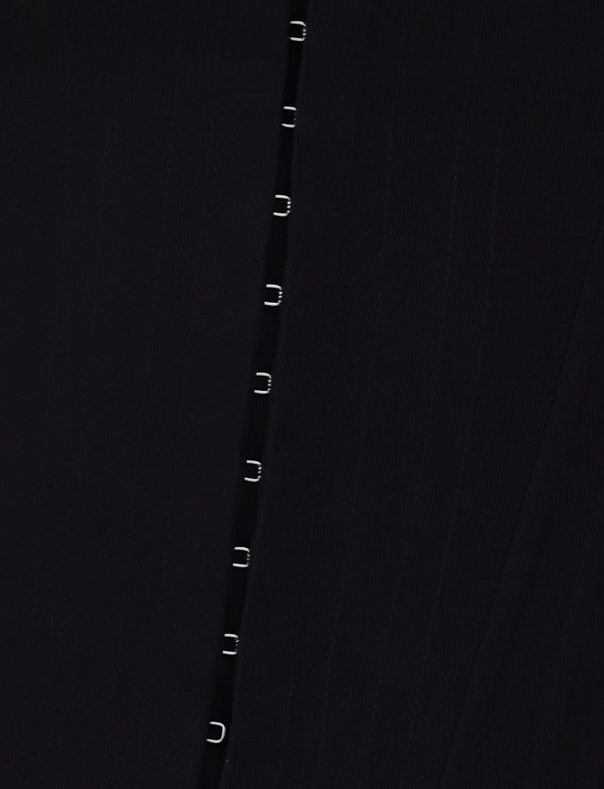 Close-up of the MILLIE CORSET 2.0 - BLACK from TIGERMIST, showcasing its textured black fabric adorned with a vertical line of evenly spaced small metallic hooks. The lighting gently accentuates the subtle shadows across the material.