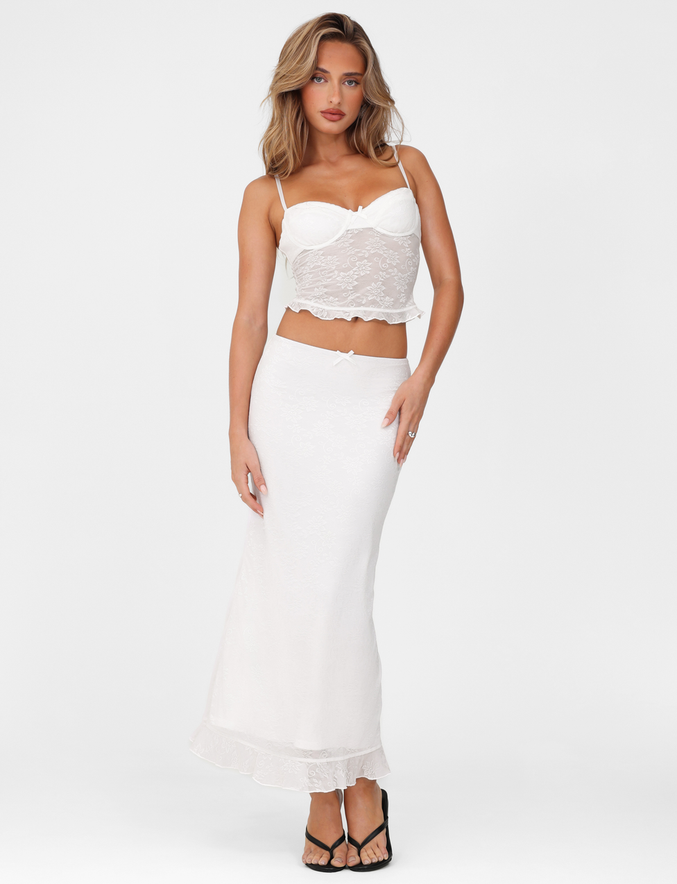 A woman models the TIGERMIST CHARLEIGH TOP – WHITE LACE, a camisole with adjustable straps, paired with a long white skirt and black sandals. She stands against a plain white background, looking at the camera with one hand on her hip.