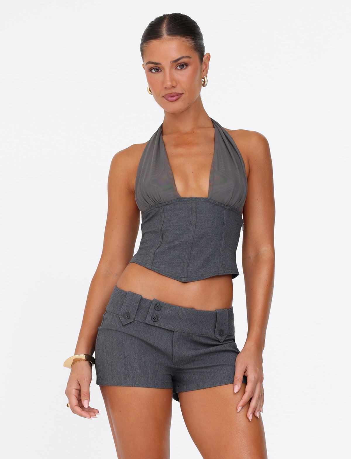 A woman stands confidently against a plain white background, wearing the JANECE TOP - GREY from TIGERMIST, crafted with grey suiting fabrication and paired perfectly with matching shorts. Her sleek hairstyle, hoop earrings, and bracelet on her left wrist enhance her poised and assured expression.