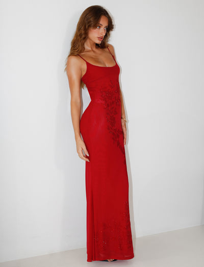 A woman in the OCTAVIA MAXI DRESS - CHERRY RED by TIGERMIST, a sophisticated long red dress with thin straps and delicate floral details, stands against a plain white background. The maxi-length piece beautifully accents her long, wavy hair as she poses with relaxed elegance.