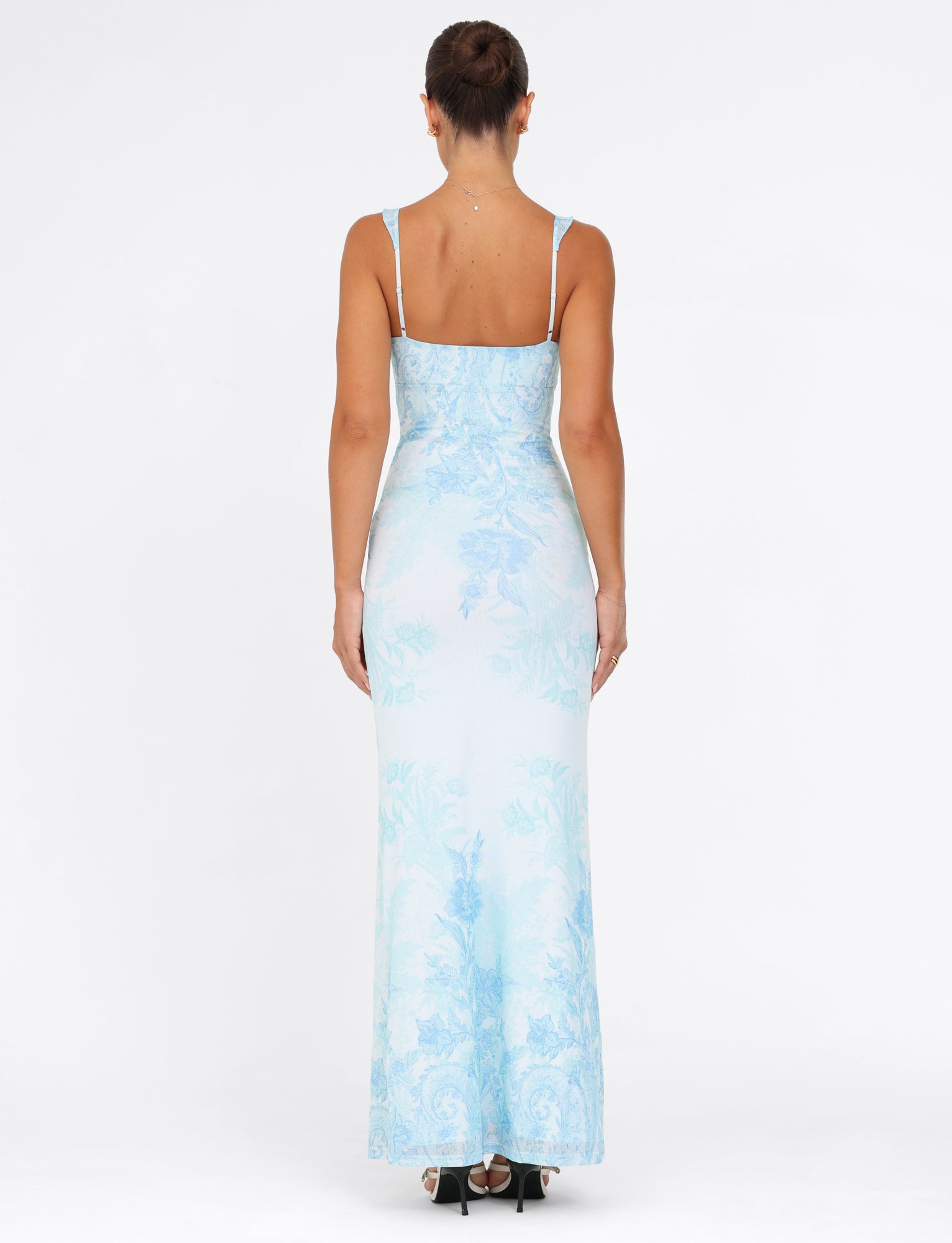 A person stands with their back to the camera, dressed in the TIGERMIST GUILIANNA FRILL MAXI DRESS - a long, blue floral tapestry dress featuring adjustable frill shoulder straps and a V-neckline. The fitted silhouette highlights their neat updo and strappy heels against a plain white background.