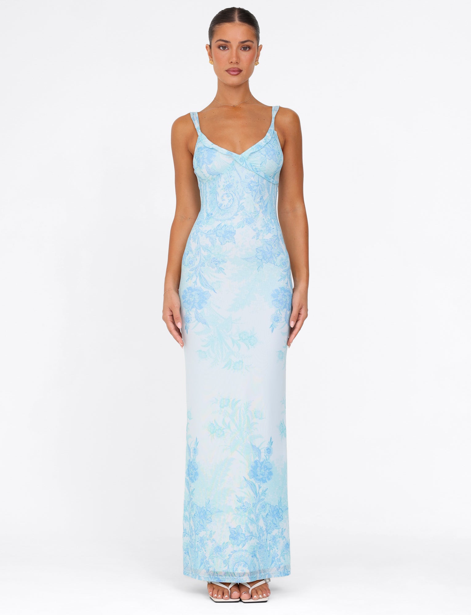 A person wearing the TIGERMIST GUILIANNA FRILL MAXI DRESS - BLUE : FLORAL : TAPESTRY stands against a plain white background. They're facing forward with their arms at their sides, showcasing the dress's elegant drape and high stretch fabric, along with its long length, light blue floral pattern, V-neck design, and adjustable frill shoulder straps.