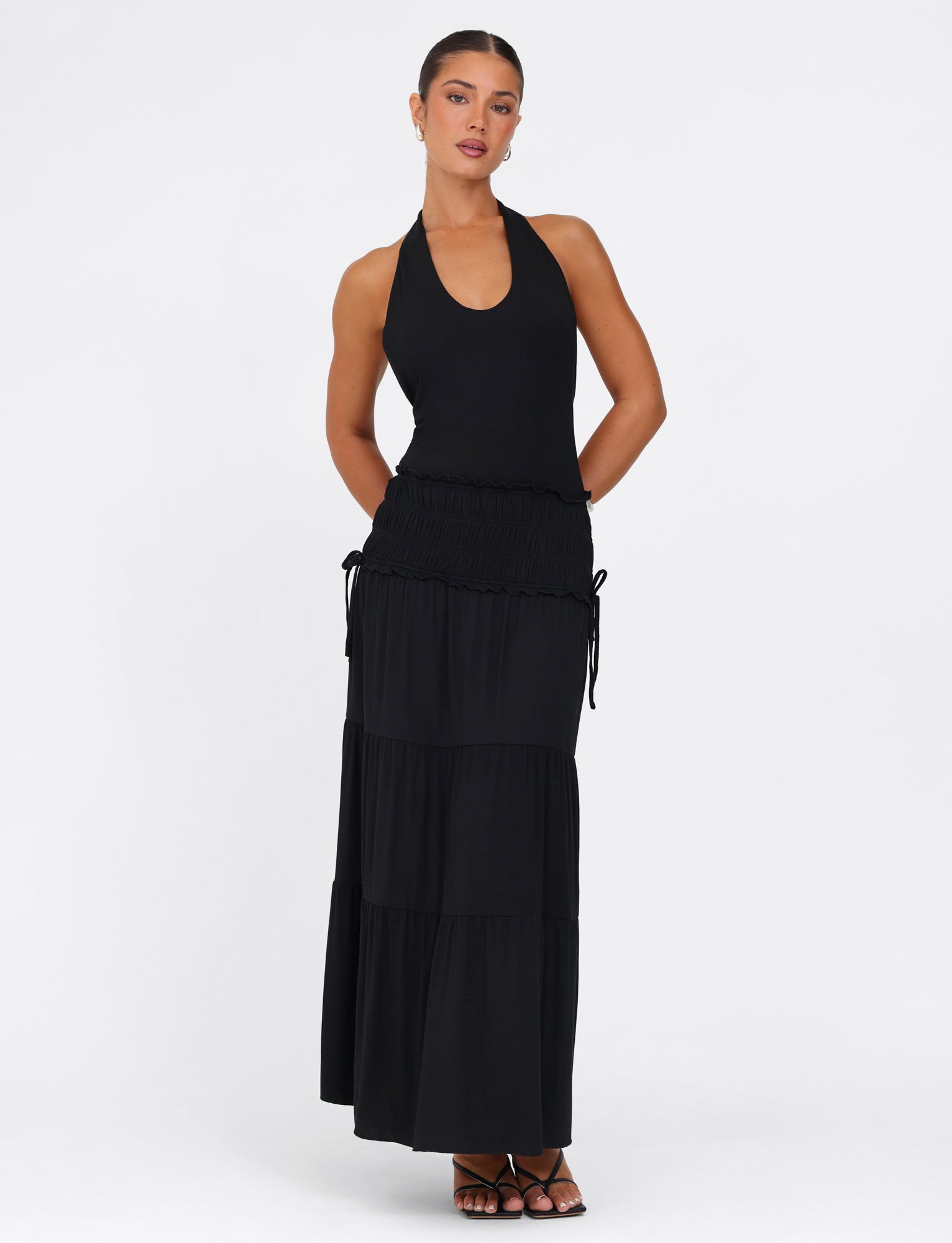 A woman models the DENALI HALTER MAXI DRESS - BLACK by TIGERMIST, standing against a plain white background. The dress is crafted from buttery jersey fabric and showcases a tiered skirt with subtle ruching at the waist. She pairs the look with black open-toed heels, keeping her arms positioned behind her back.