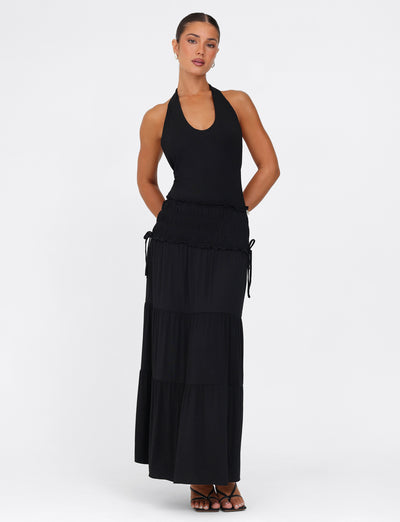 A woman models the DENALI HALTER MAXI DRESS - BLACK by TIGERMIST, standing against a plain white background. The dress is crafted from buttery jersey fabric and showcases a tiered skirt with subtle ruching at the waist. She pairs the look with black open-toed heels, keeping her arms positioned behind her back.