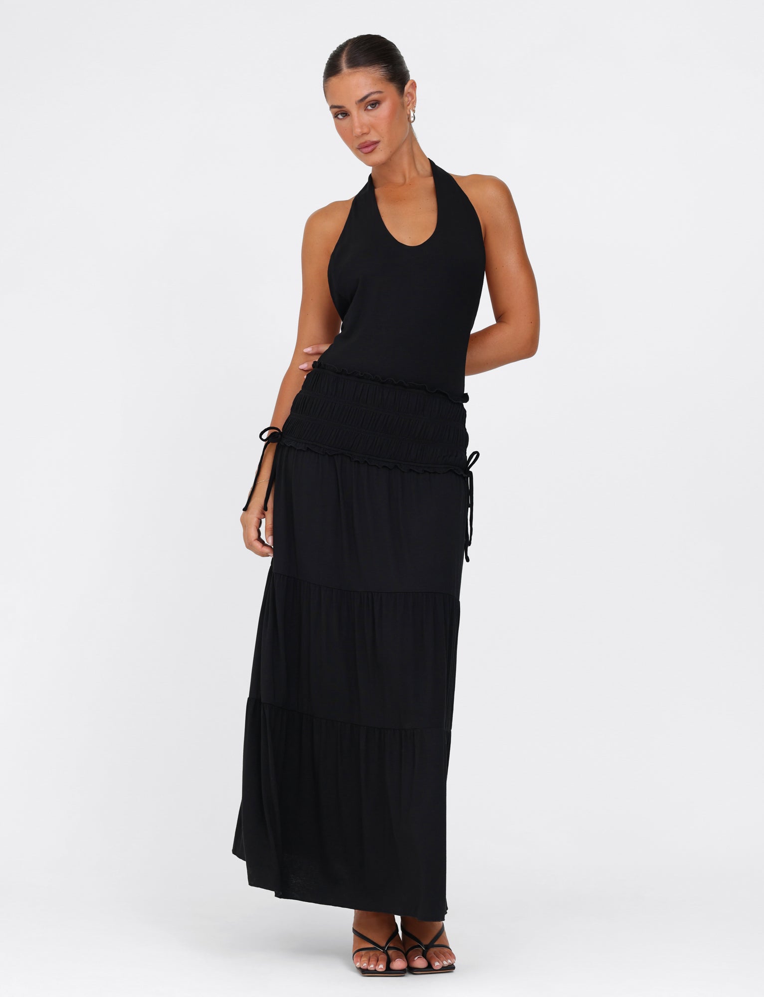 A woman in the TIGERMIST DENALI Halter Maxi Dress - Black stands against a plain backdrop, showcasing the dress's elegant tiered skirt. Her hair is styled in an elegant updo, and she adds flair to her chic ensemble with earrings and sandals. The dress's low back enhances the allure of her look.