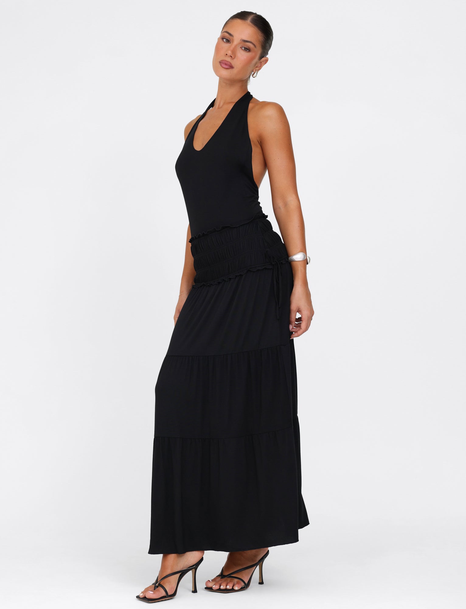 A woman poses elegantly against a white backdrop in the DENALI HALTER MAXI DRESS - BLACK by TIGERMIST. The sleeveless dress, with its halter neck and low back design, adds sophistication as she stands with one hand on her hip. Complementing her look are black high-heeled sandals, an eye-catching bracelet, and an elegant updo.