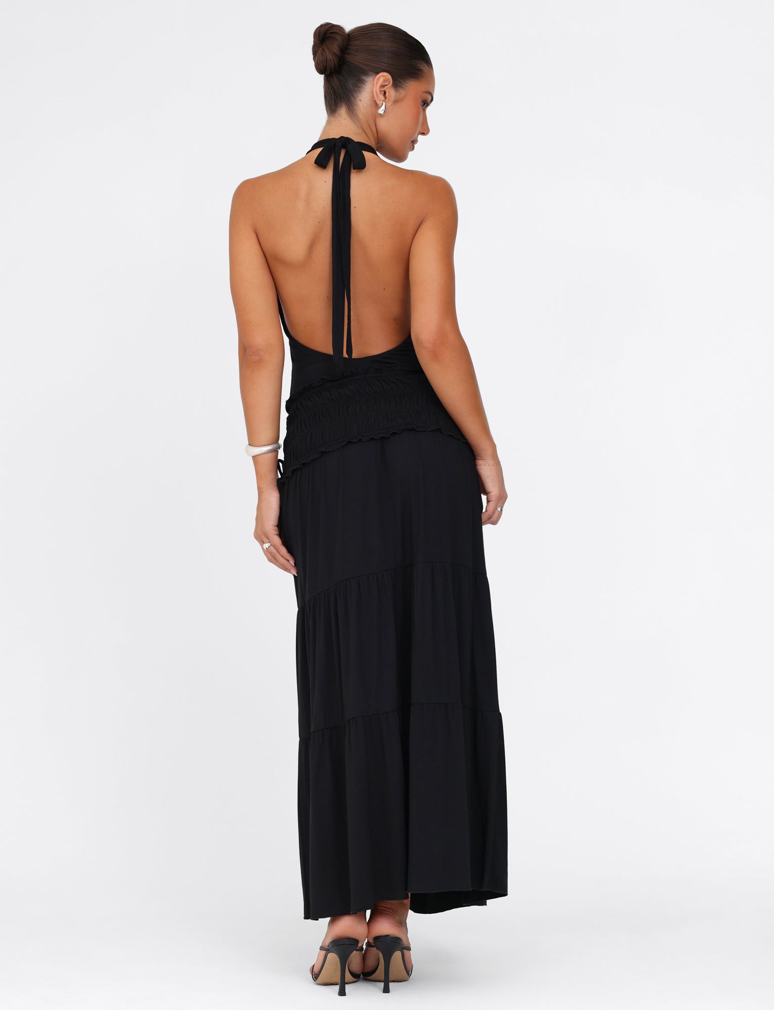 A woman models the DENALI HALTER MAXI DRESS in classic black from TIGERMIST, featuring a buttery jersey fabric. The dress is sleeveless and backless with a halter neck. She completes her look with heels and has styled her hair in a sleek bun, all set against a plain white background.