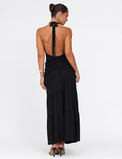 A woman models the DENALI HALTER MAXI DRESS in classic black from TIGERMIST, featuring a buttery jersey fabric. The dress is sleeveless and backless with a halter neck. She completes her look with heels and has styled her hair in a sleek bun, all set against a plain white background.