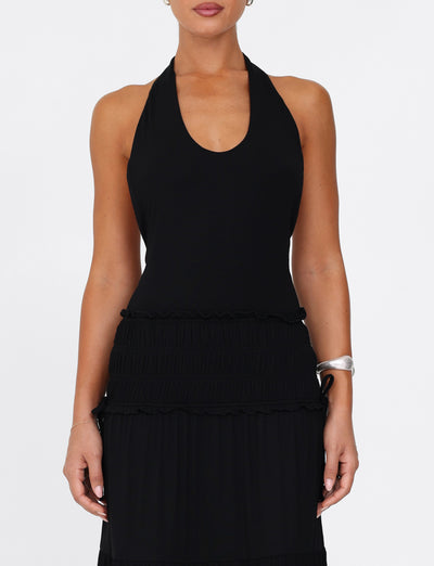 A person wearing the DENALI HALTER MAXI DRESS by TIGERMIST in black stands against a plain background. A silver cuff bracelet adorns their right wrist. The dress features a fitted bodice and a wavy, tiered skirt with textured detailing, creating an elegant yet modern look.