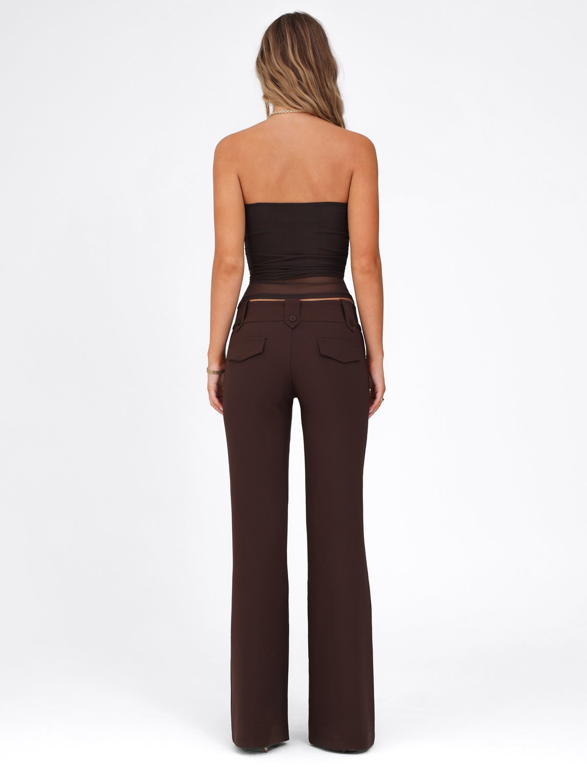 A woman faces away, wearing a black strapless top and TIGERMIST KITTIE PANT - BROWN : CHOCOLATE, a high-waisted flare with back pockets and an asymmetrical waistband, set against a plain white background.