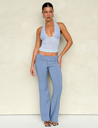 The individual stands against a plain background, dressed in a light blue halter top and the KITTIE PANT - BLUE in steel blue by TIGERMIST. The look is enhanced with black strappy heels, sleek slicked-back hair, and hoop earrings.