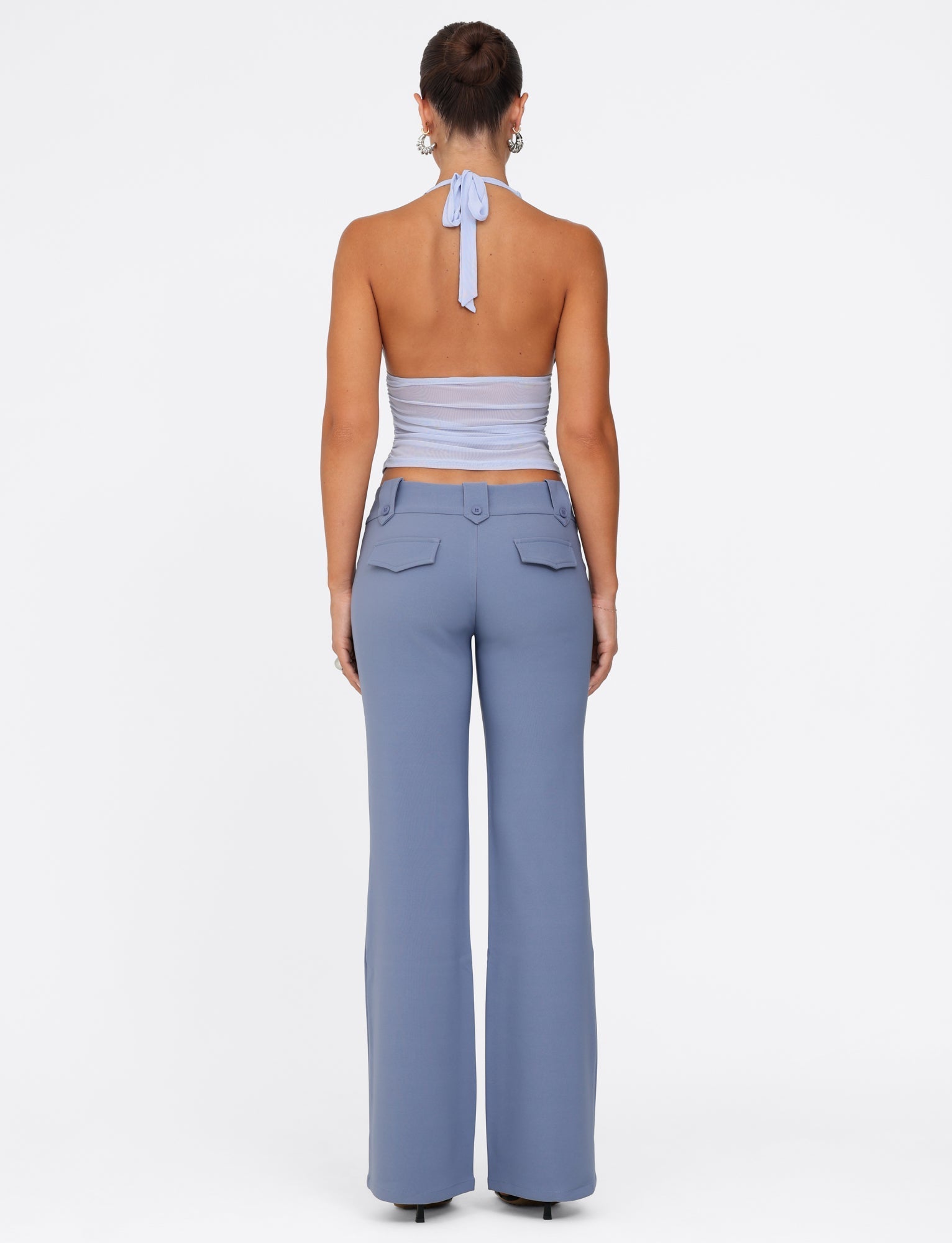 A person stands facing away, dressed in the light blue halter crop top paired with TIGERMIST's KITTIE PANT - BLUE in steel blue. Their hair is tied up and accented by hoop earrings, all set against a plain white background.