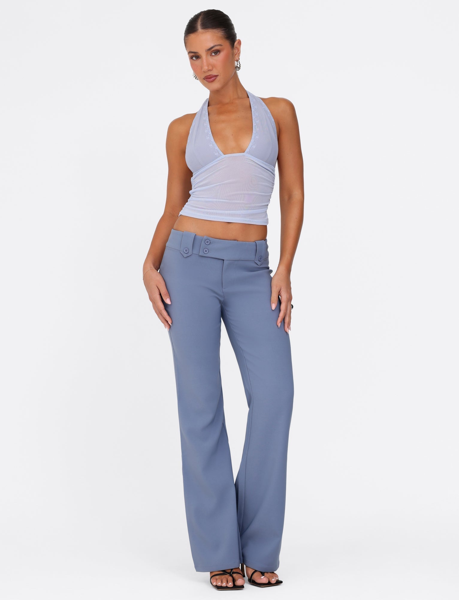 A person poses confidently against a white background, wearing a sleeveless, halter-neck lavender top paired with the KITTIE PANT in steel blue by TIGERMIST. They accessorize with hoop earrings and black heeled sandals.