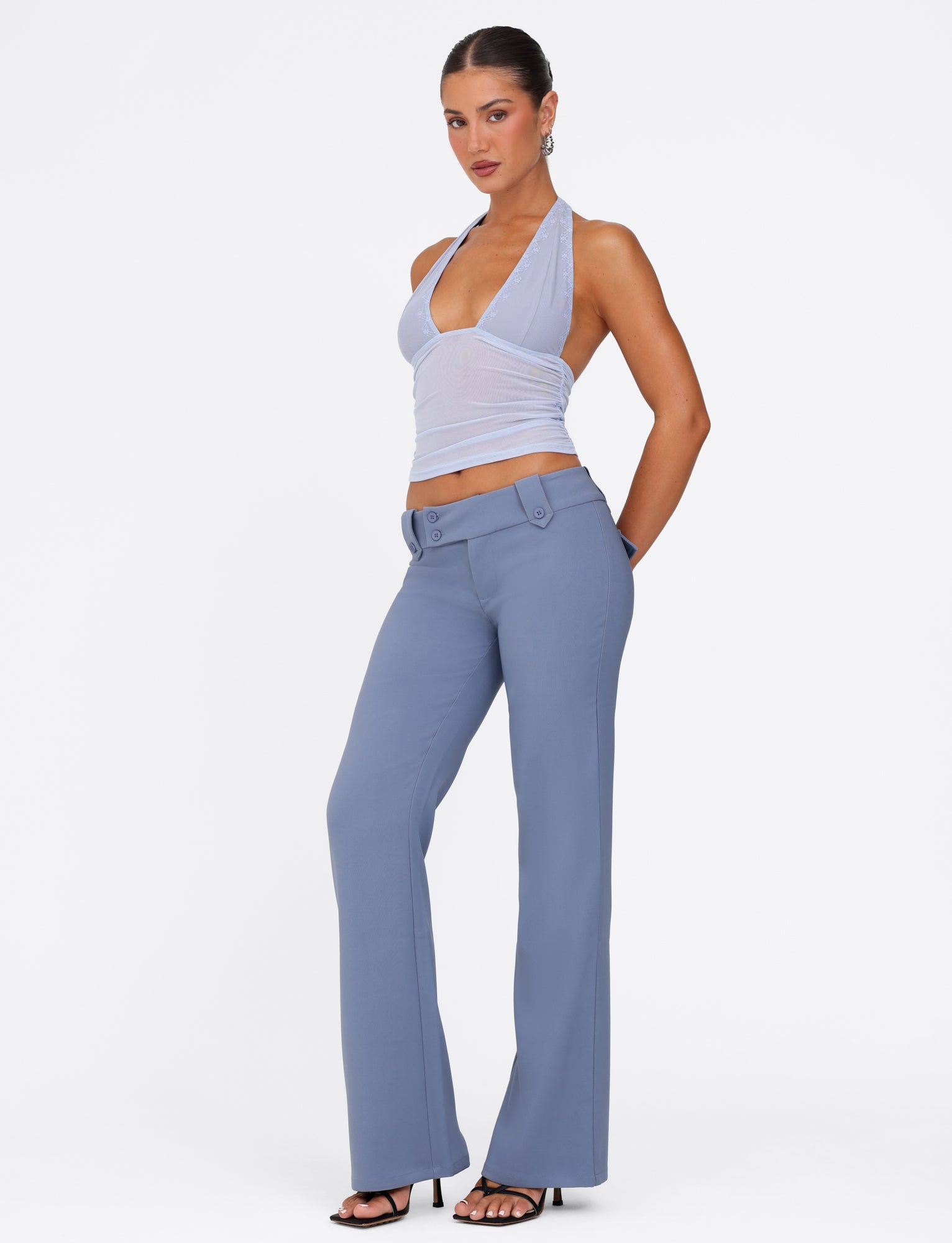 A person stands confidently in the trendy "KITTIE PANT - BLUE" from TIGERMIST, paired with a light blue halter top and complemented by black high heels. The plain white background elegantly highlights this stylish ensemble.