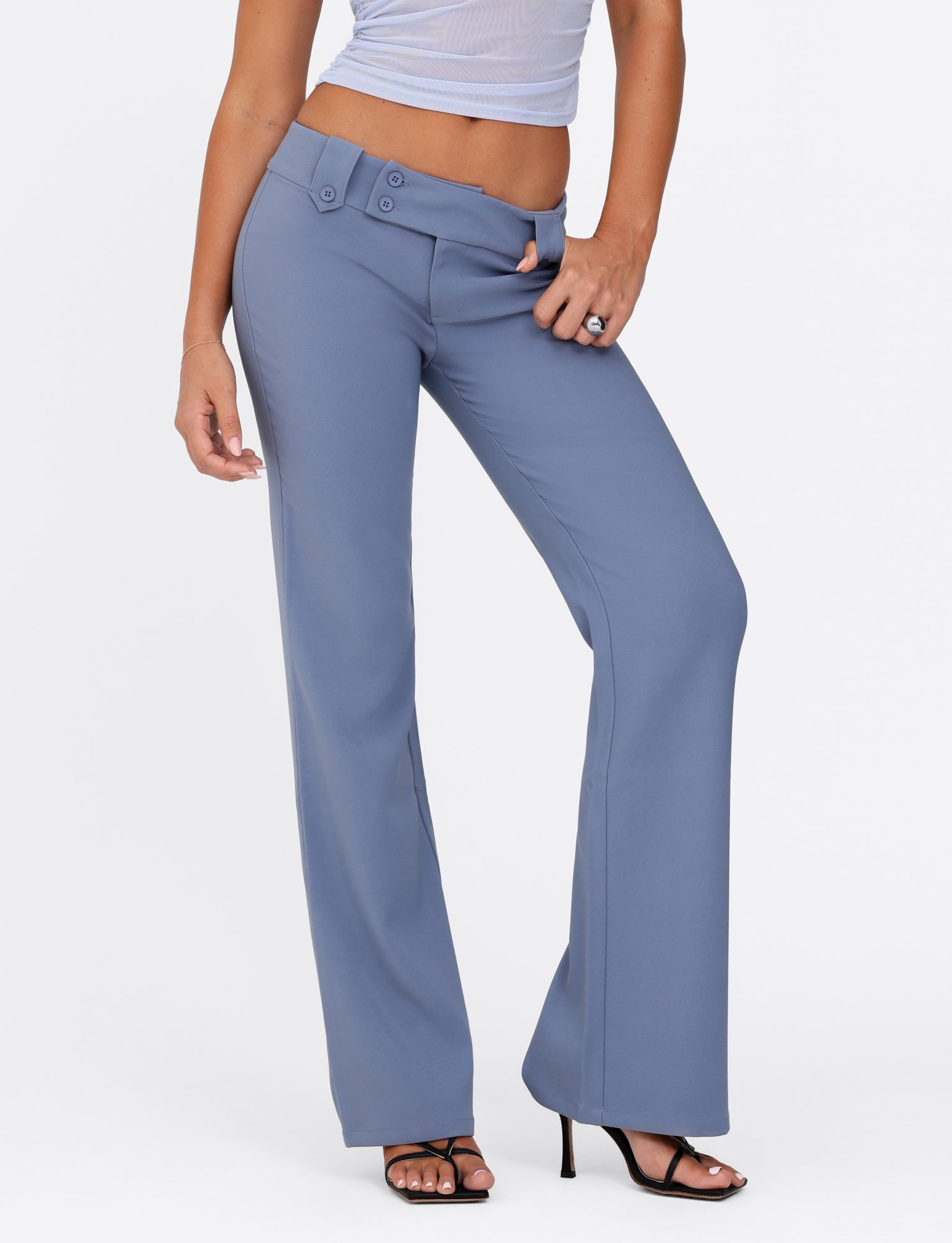 A person dressed in the KITTIE PANT - BLUE from TIGERMIST, featuring a steel blue color and flare fit with button details at the waist, paired with a sheer white top. They are standing with one hand on their hip, complementing their outfit with black high-heeled sandals.