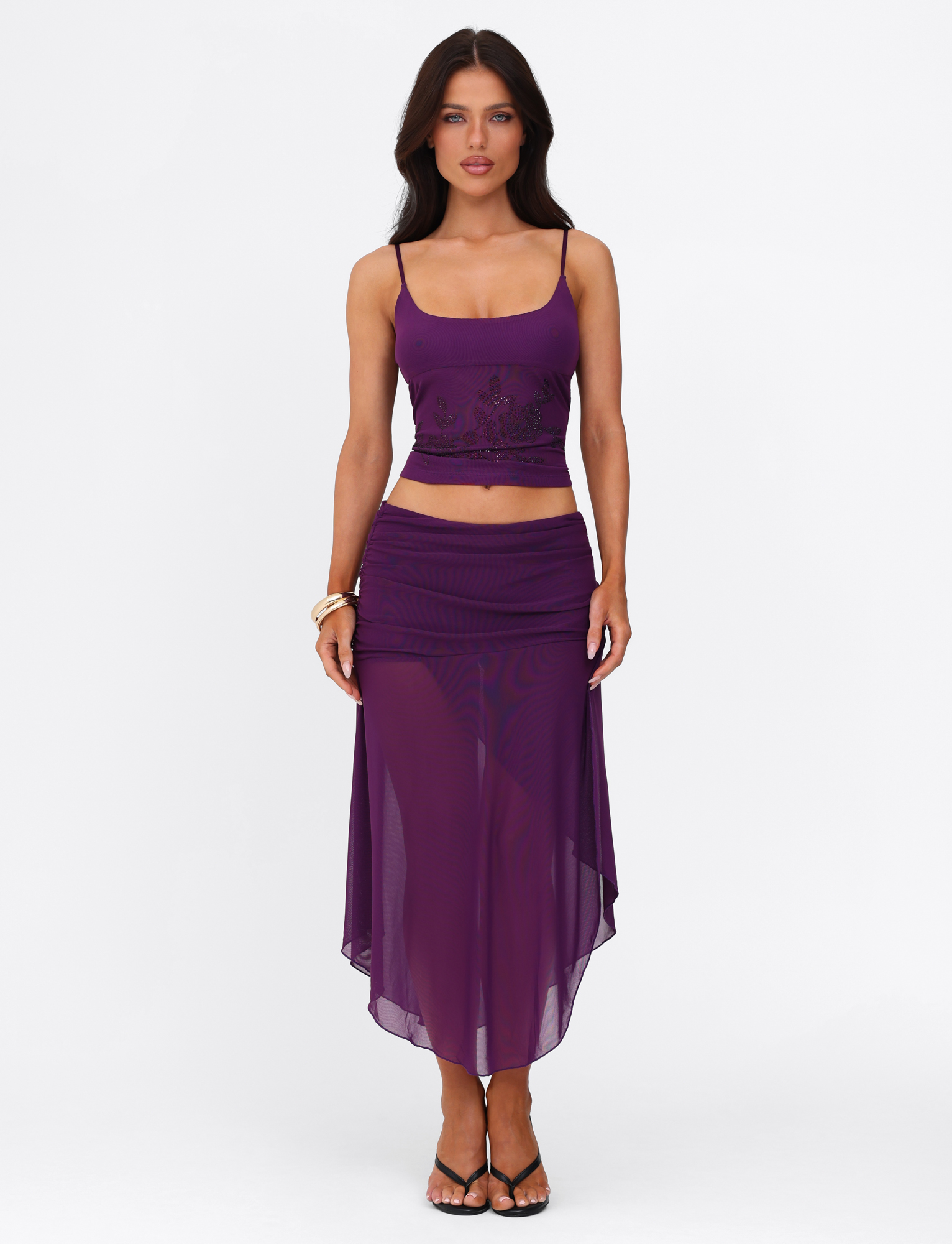 A woman with long brown hair wears the TIGERMIST OCTAVIA TOP in purple against a plain white background, paired with a sheer, flowing purple skirt, high heels, and gold bangles on her right wrist.
