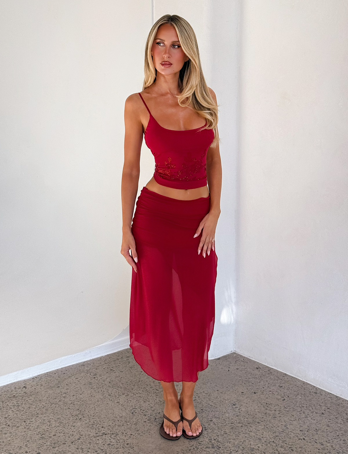 A woman with long blonde hair wears the TIGERMIST OCTAVIA TOP - RED and a matching sheer floral skirt, standing indoors by a white wall and looking to the side. She completes her look with black sandals.
