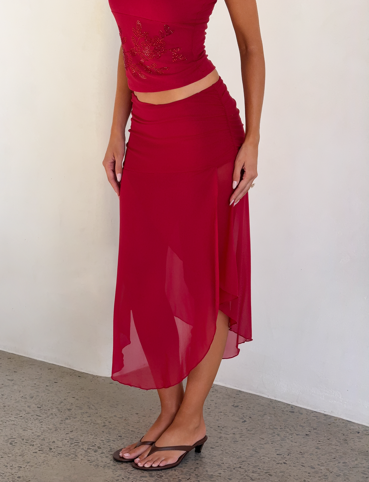 A woman wears the TIGERMIST MARIANA SKIRT - RED, featuring a ruched waistband and sheer midi length, styled with a sleeveless red beaded top and black open-toe heels. She stands against a white wall, her face not shown.