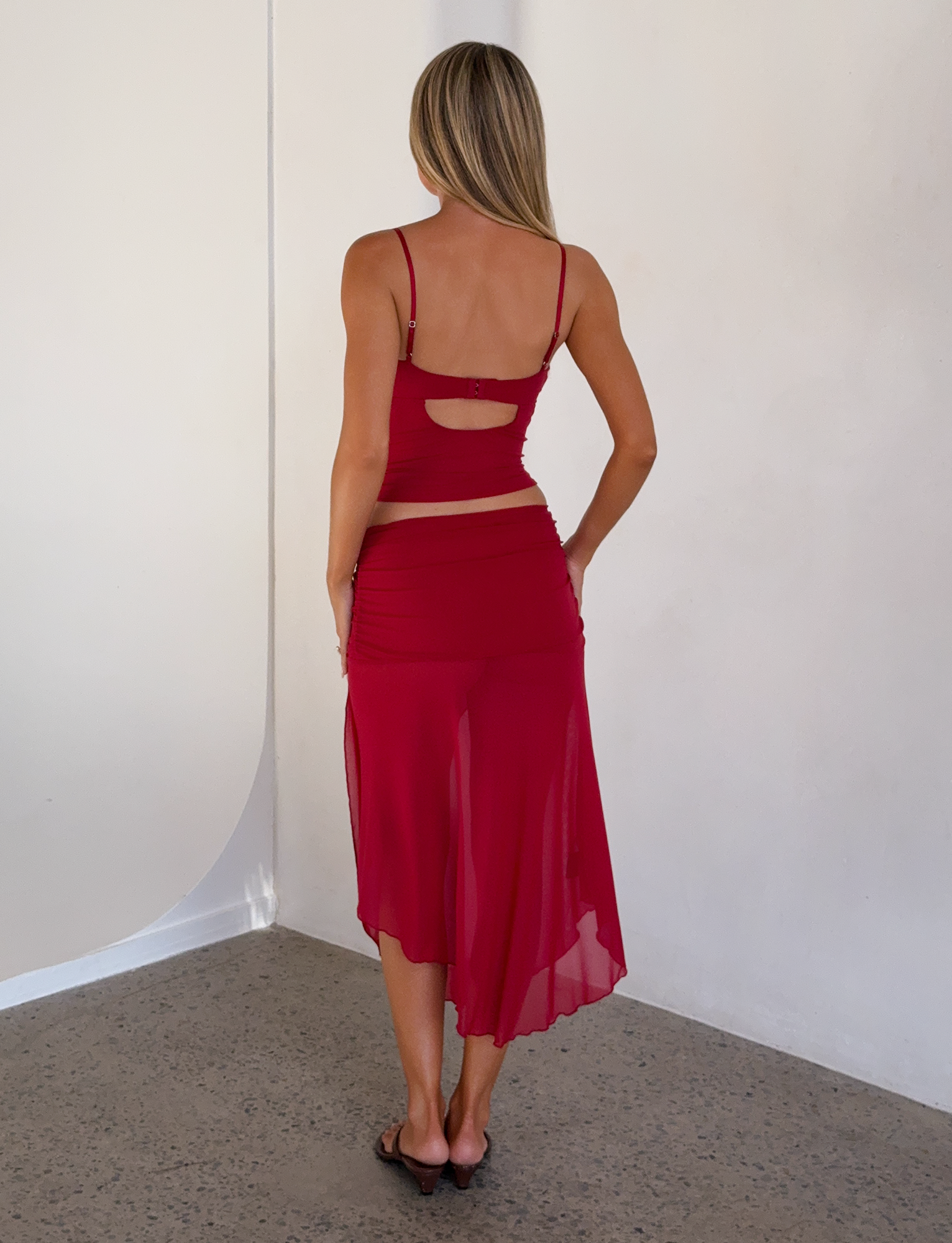 A woman with long blonde hair, barefoot on a speckled floor against a white wall, wears a sleeveless red top and the TIGERMIST MARIANA SKIRT - RED featuring a sheer design and ruched waistband.