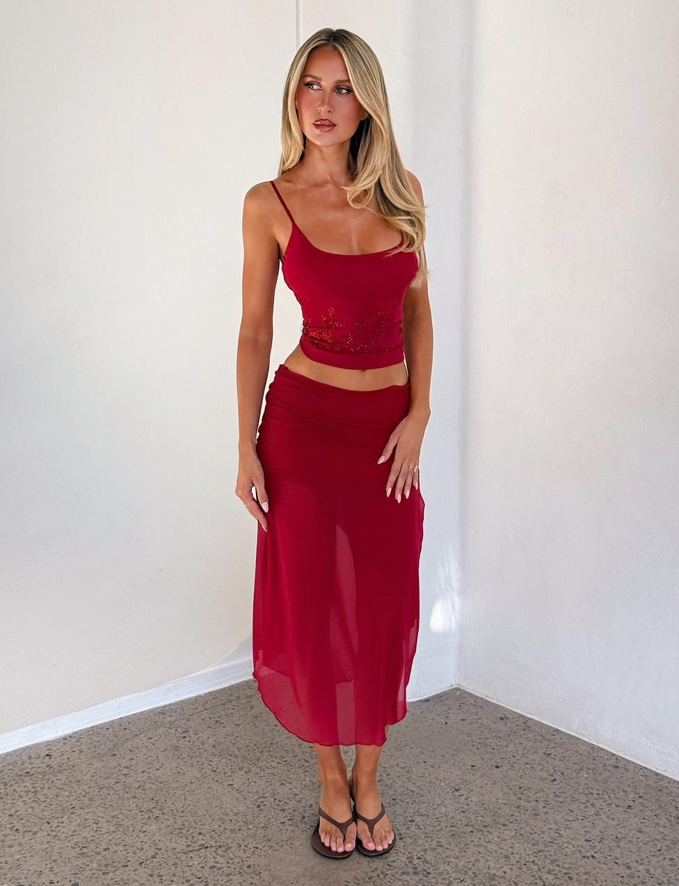 A woman with long blonde hair wears the CL - MARIANA SKIRT by Tiger Mist, styled with a red fitted crop top. She stands indoors against a white wall on a gray floor, looking to the side with one hand by her leg.