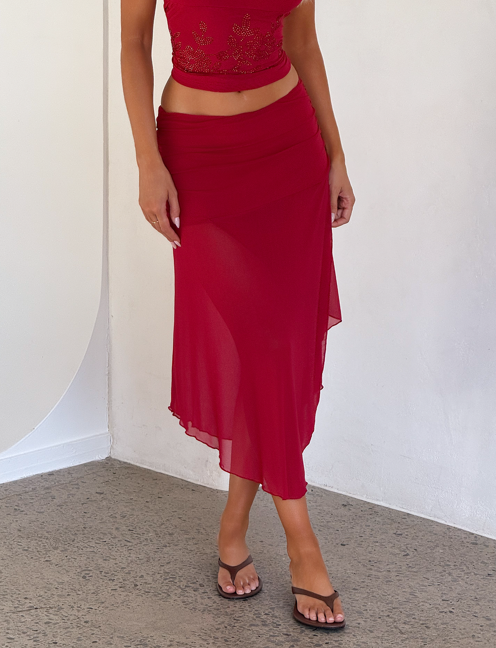 A person in a sleeveless, embroidered red crop top and the TIGERMIST MARIANA SKIRT - RED, with a ruched waistband and asymmetric wrap detail, stands against a white wall in tan sandals, left hand resting by their side.
