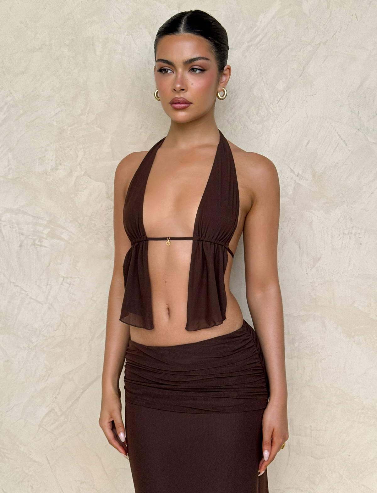 A woman in the TIGERMIST ESHA TOP - BROWN : CHOCOLATE with adjustable ties and a matching skirt stands against a beige textured wall, wearing gold hoop earrings and slicked-back hair, looking to the side with a neutral expression.