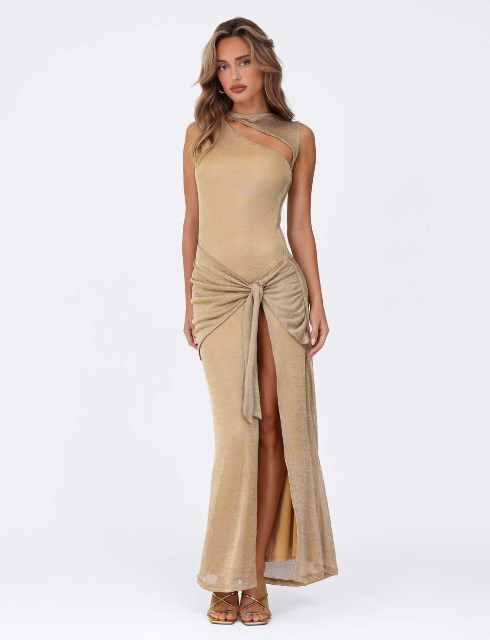 A woman in the TIGERMIST AZELMA MAXI DRESS - GOLD, a sleeveless metallic knit maxi with a neckline side cutout and front tie, poses against a white background. She has wavy hair and wears strappy sandals.