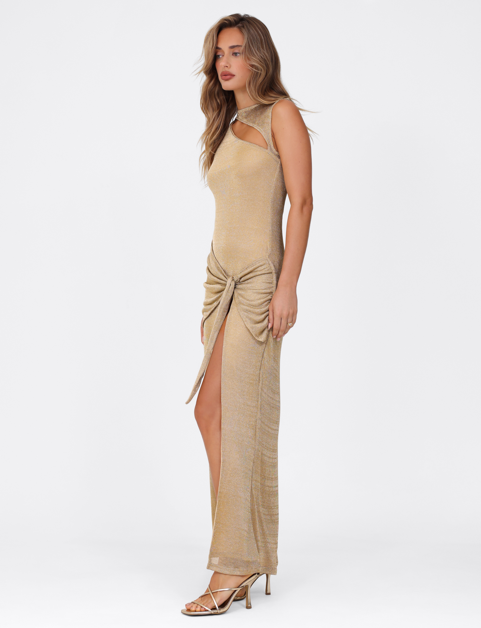 A woman in profile wears the TIGERMIST AZELMA MAXI DRESS - GOLD, a metallic knit maxi with a neckline side cutout, ruched knot at the hip, high slit, and strappy high heels. She has long wavy hair and a neutral expression.