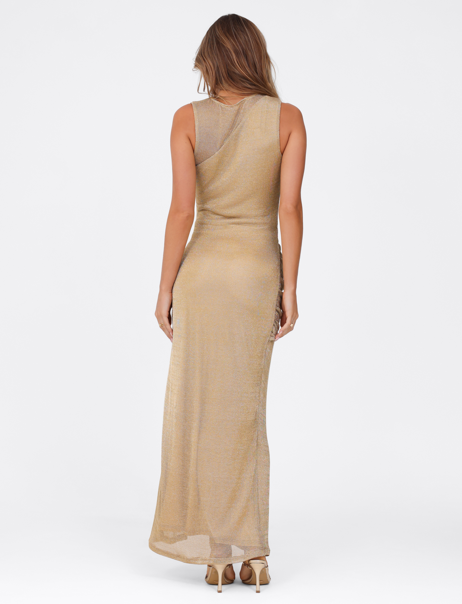 A woman with light brown hair and beige heels faces away, wearing the TIGERMIST AZELMA MAXI DRESS - GOLD, a sleeveless metallic knit maxi dress with an asymmetrical neckline, set against a plain white background.
