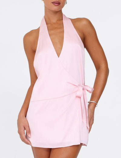 The individual is stylishly attired in a pastel pink Monroe 2.0 Mini Dress by TIGERMIST, showcasing a satin wrap style with an adorable bow accent at the waist. A minimalist bracelet flawlessly completes this sophisticated ensemble.