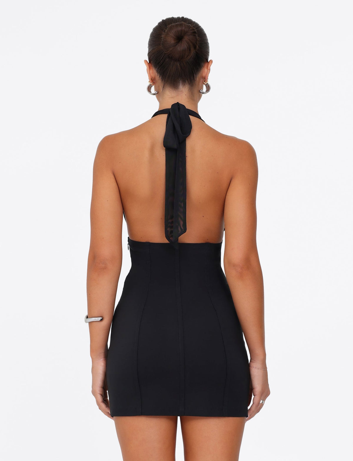 A woman stands with her back to the camera, wearing the JANECE MINI DRESS - BLACK by TIGERMIST. Her hair is styled in a bun, and she accessorizes with hoop earrings. She holds one arm by her side while the other is bent, all set against a plain white background that accentuates the dress's elegance and form-fitting design.