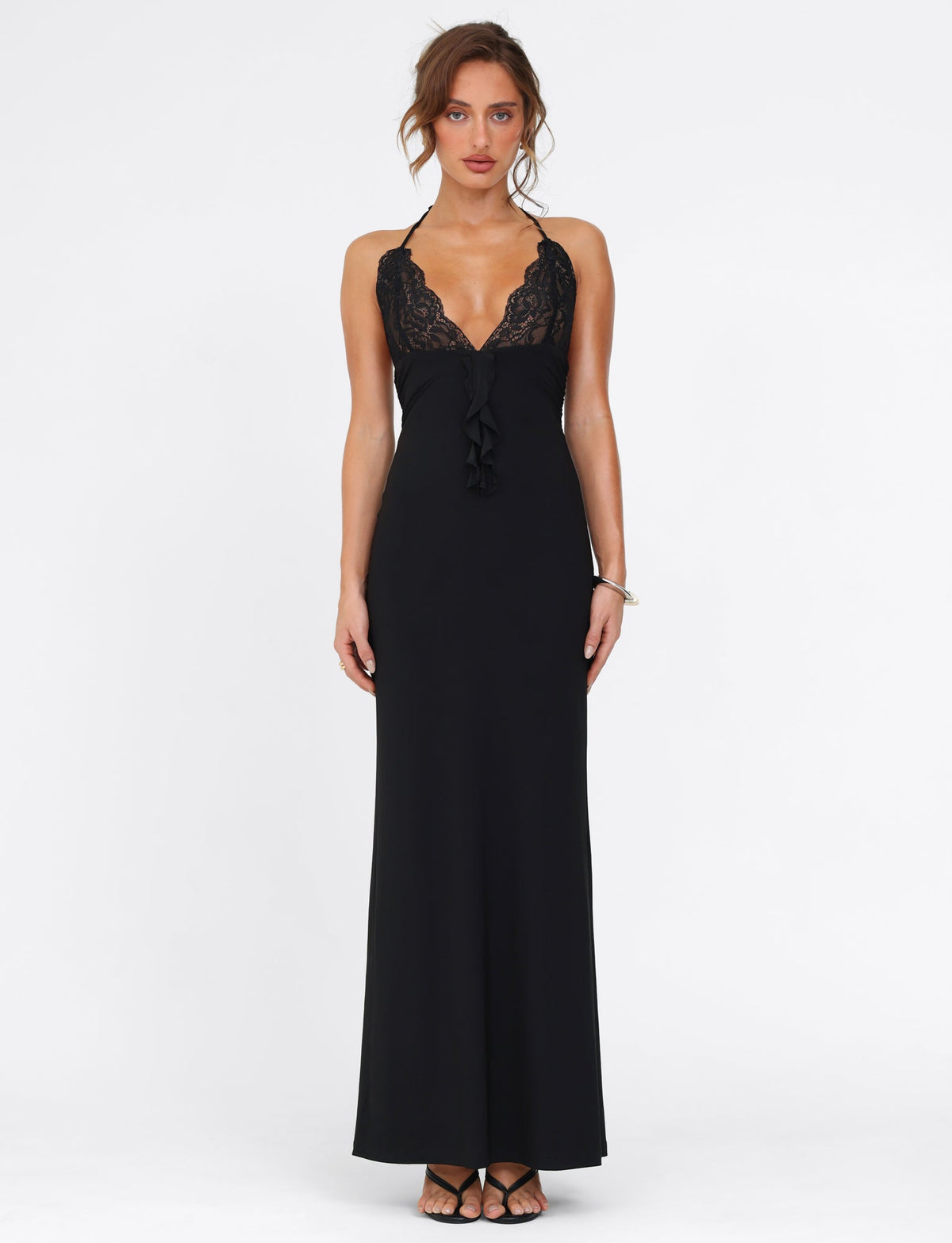 A woman stands against a white background wearing the DEENA MAXI DRESS by TIGERMIST. This elegant black dress features lace bust cups that beautifully enhance the neckline. Her hair is tied up, and she completes her look with black shoes and a silver bracelet, exuding timeless sophistication.