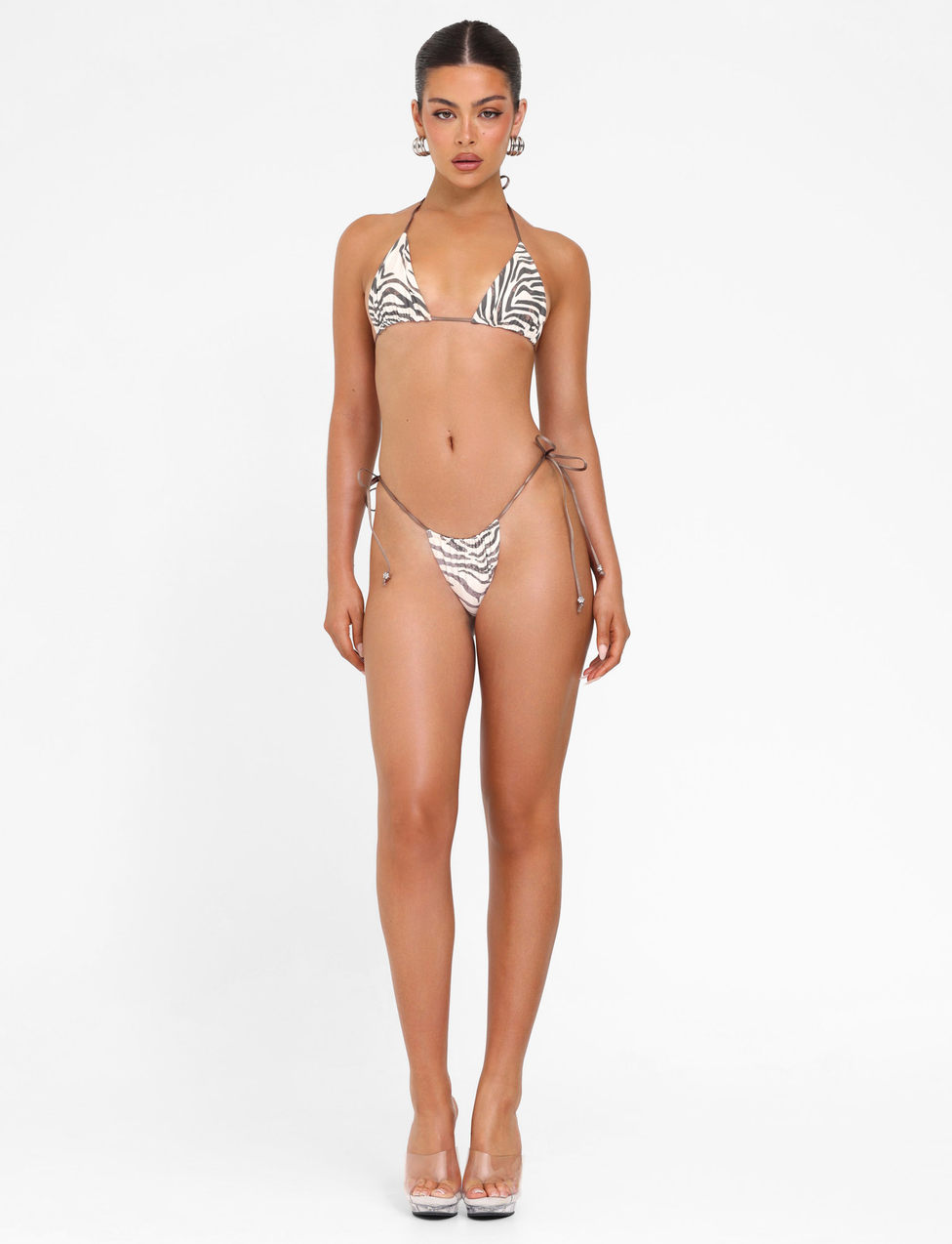 A woman faces forward wearing the TIGERMIST AVE BIKINI TOP - NEUTRAL : ZEBRA, a true-tie halter neck, no padding triangle bikini top, paired with clear high heels against a plain white background.