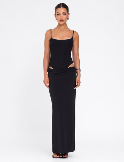 A woman faces forward in the TIGERMIST UMA DRESS - BLACK, a bodycon maxi with adjustable spaghetti straps and cut-out hip details. Her hair is pulled back; she accessorizes with hoop earrings and bracelets, against a plain white background.